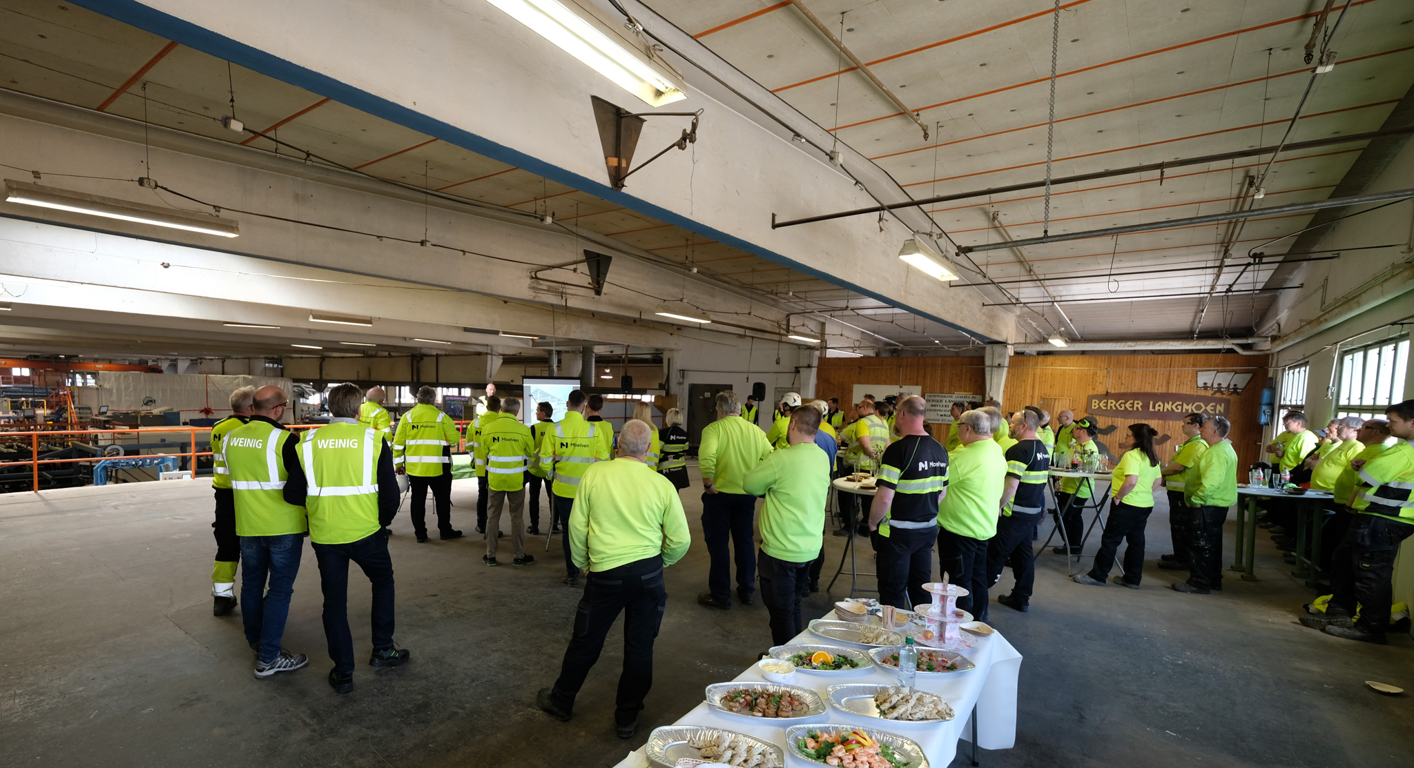 Ansatte i Moelven Langmoen som  står med ryggen til i stor hall. Foto