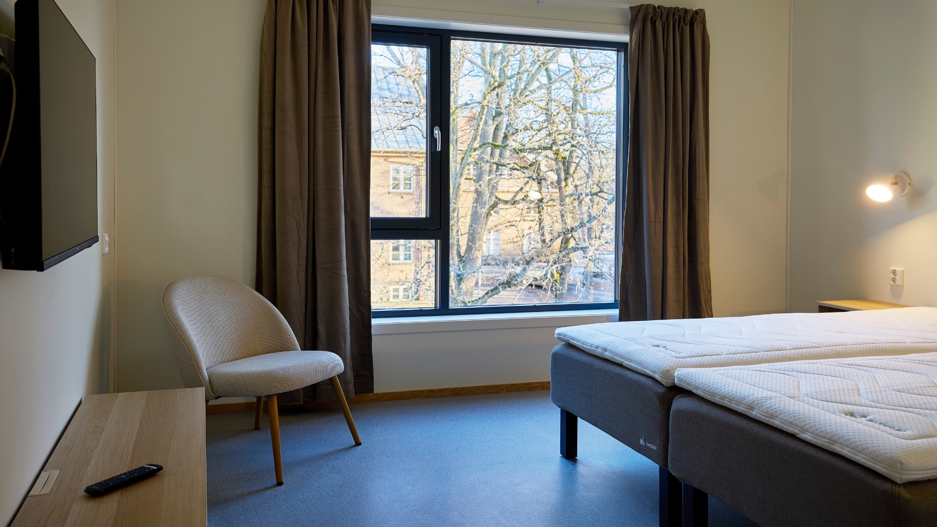Hotellrom med dobbeltseng, TV-benk og perspektiv mot vindu.