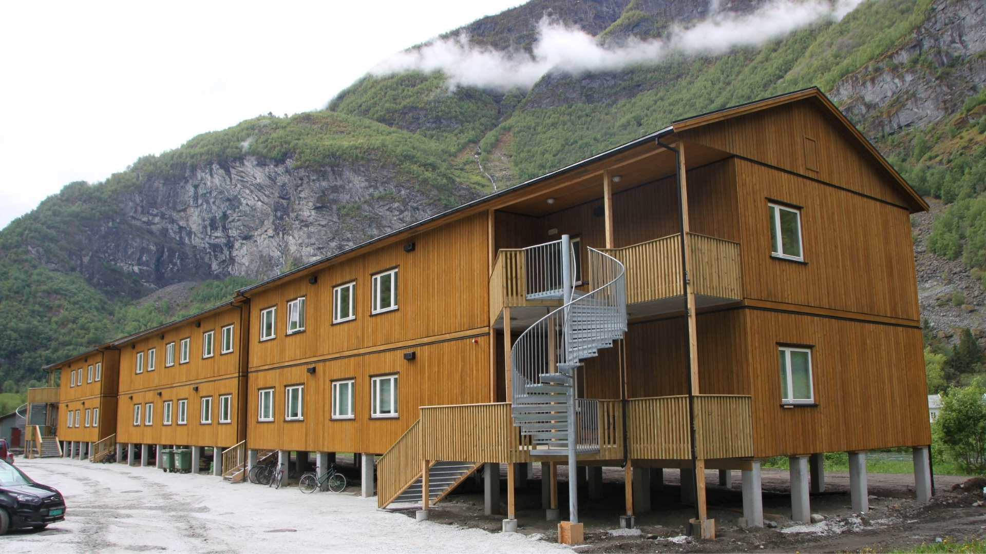 Et langt bygg med lys brun trekledning står nedenfor et bratt fjell.