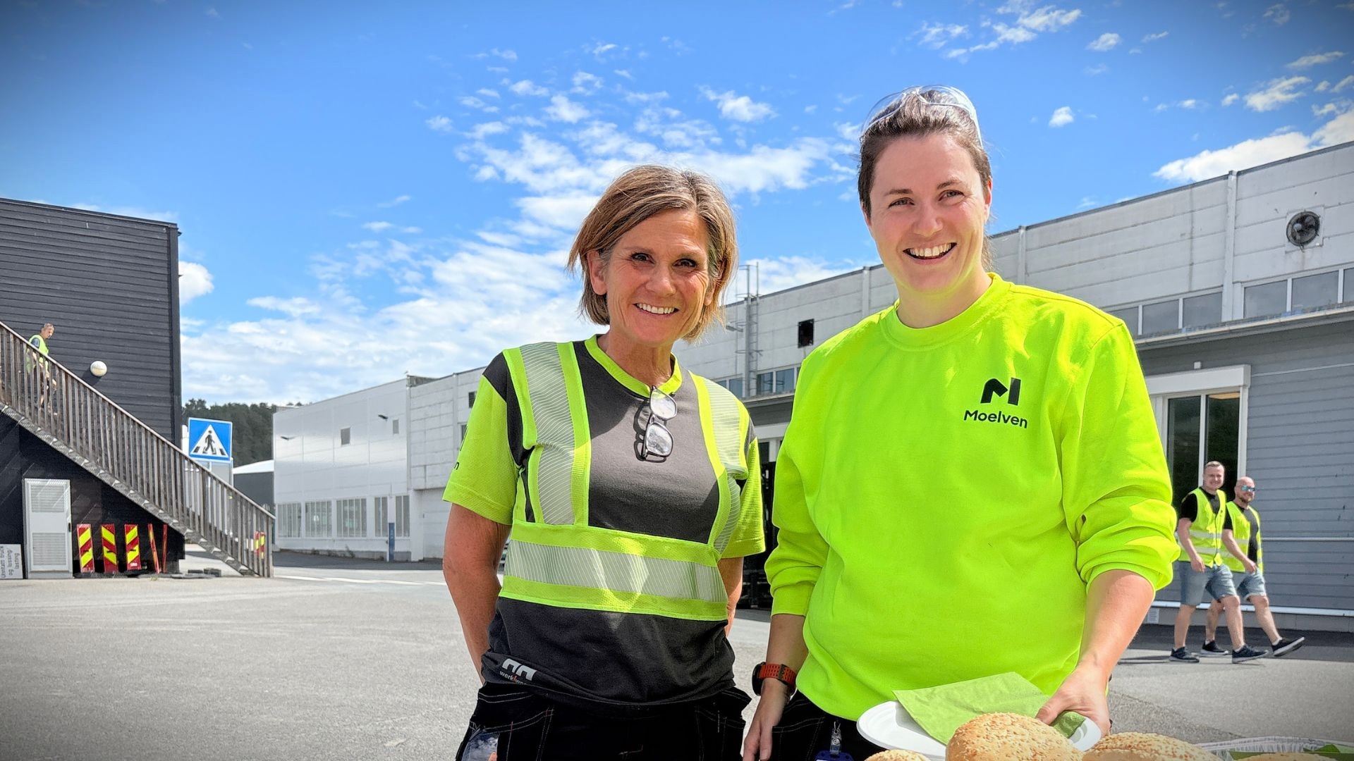 To smilende kvinner i synlighetstøy utenfor Mo9elven Byggmodul sine fabrikklokaler på Moelv.