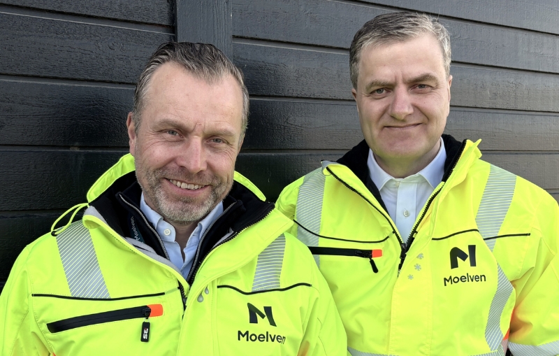 To smilende menn i Moelven synlighetstøy står inntil en brunmalt vegg.