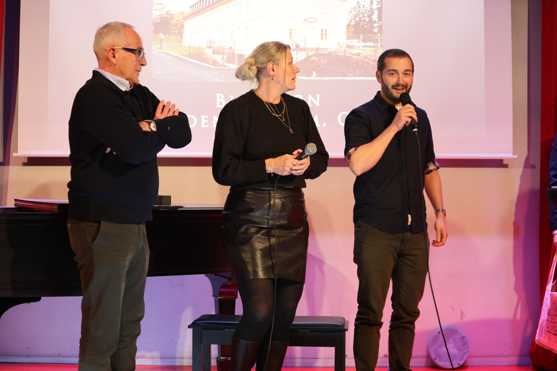 Tre personer står på en scene. Den ene personen holoder en mikrofon.