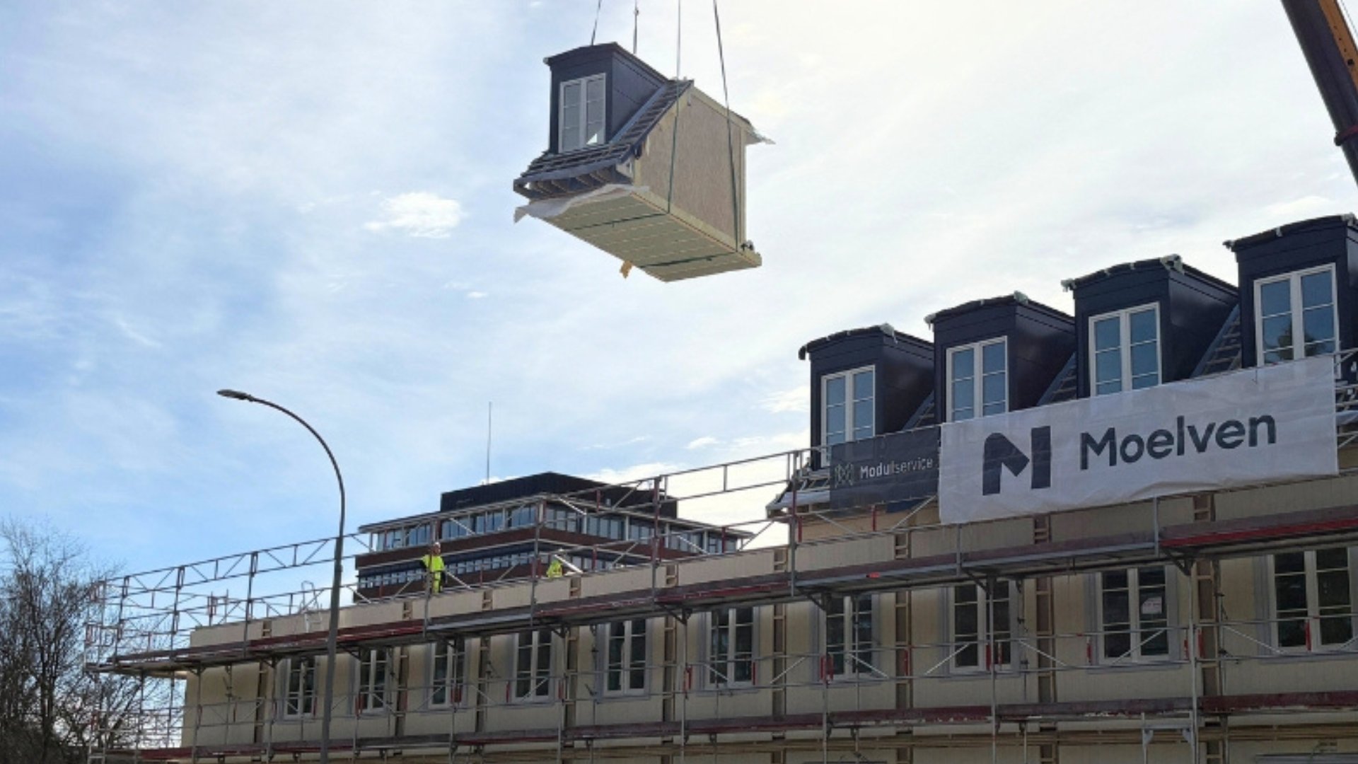 Ein Holzmodul, das an einem Kran in der Luft hängt, über einer Modulbaustelle.