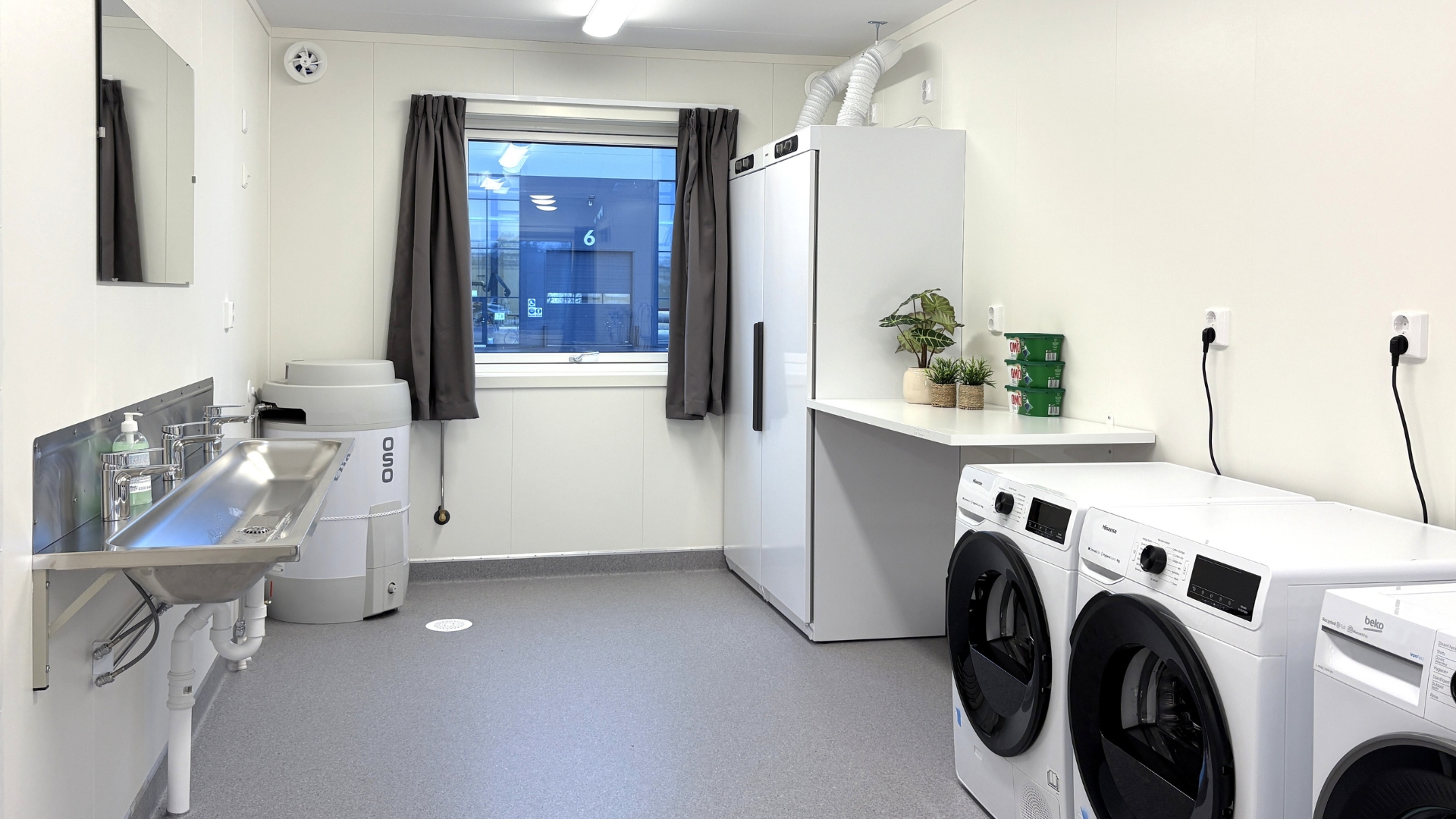 Fælles vaskerum i boligpavillon med vaskemaskiner, tørreskab og vask til arbejdere.