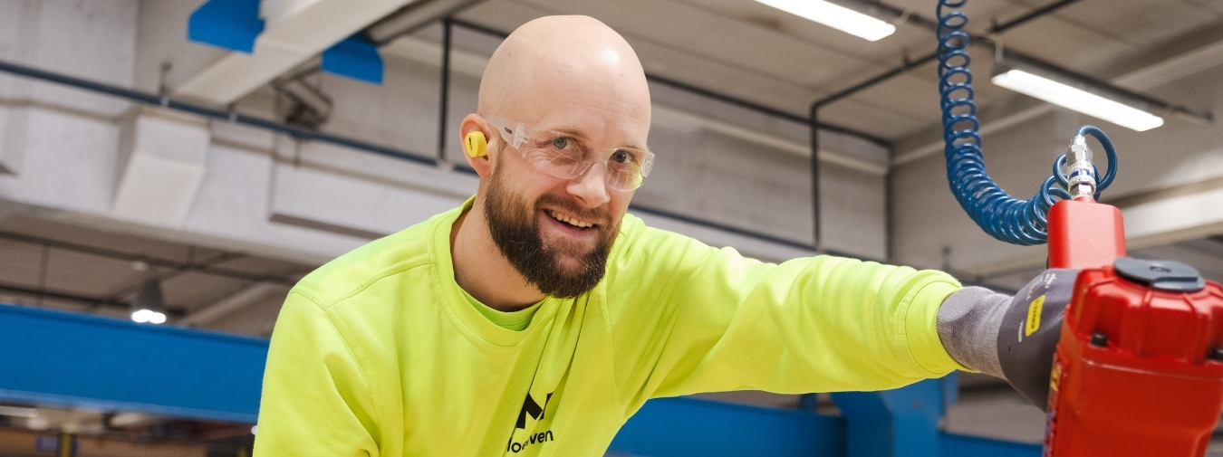 Smiling man in a factory looks at the camera and holds a nail gun.