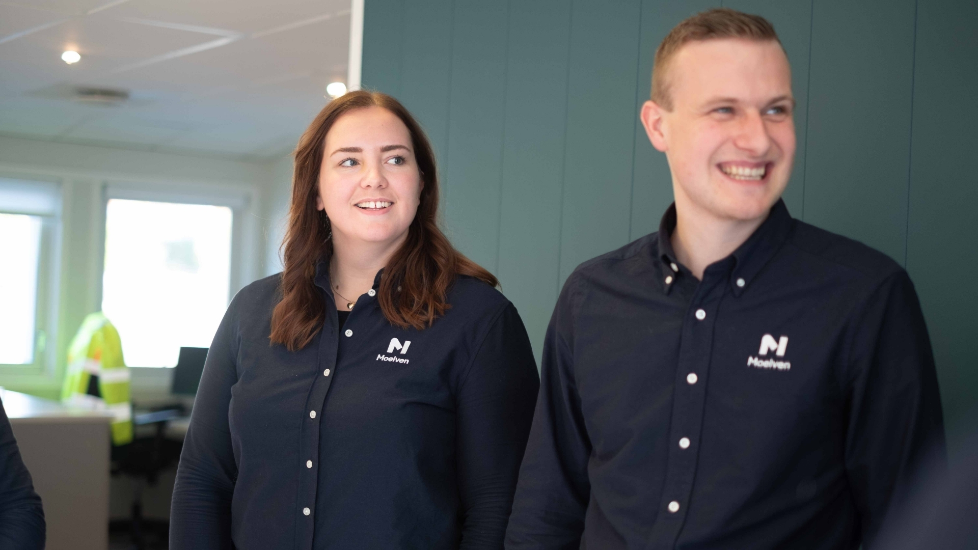 Brosandi kona og maður í bláum Moelven-skyrtum horfa til vinstri.