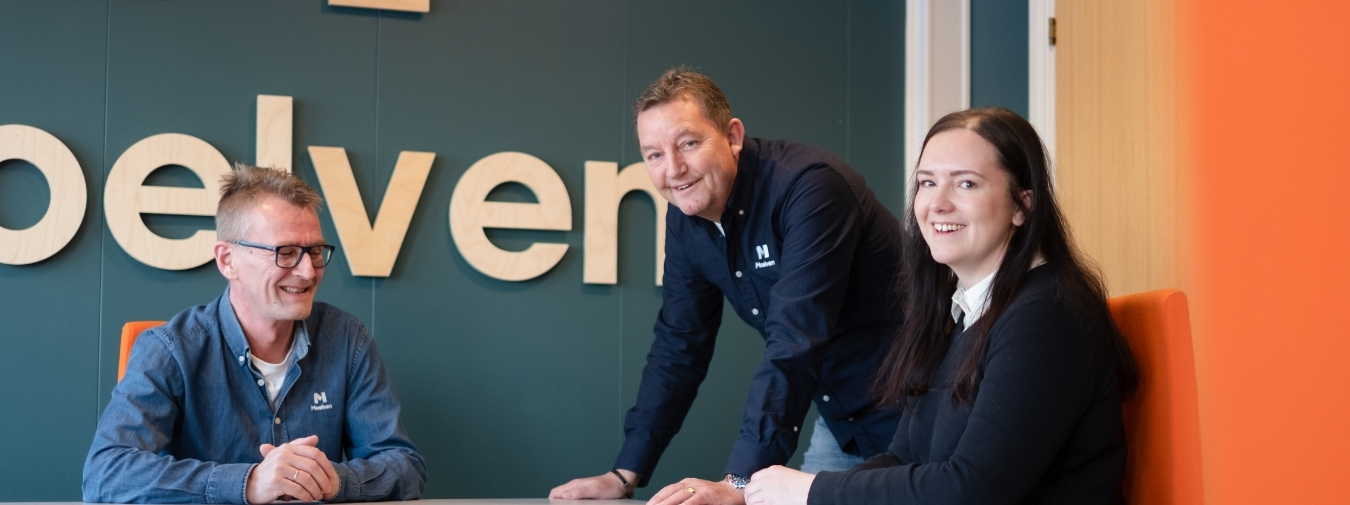 Þrír brosandi samstarfsmenn sitja við borð í fundarherbergi.