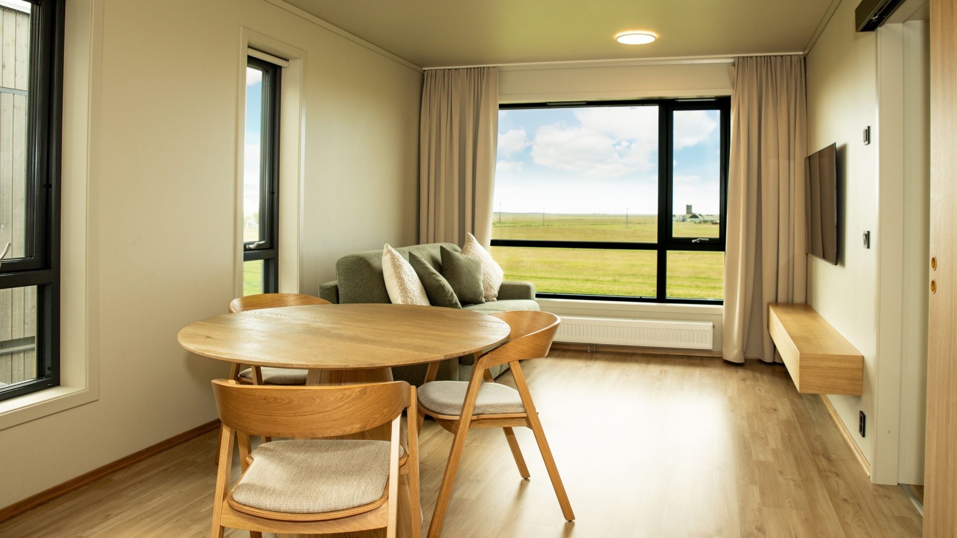 Björt setustofa í modúlbyggðu hótelherbergi með borðstofuborði og stórum gluggum með útsýni yfir opið landslag.