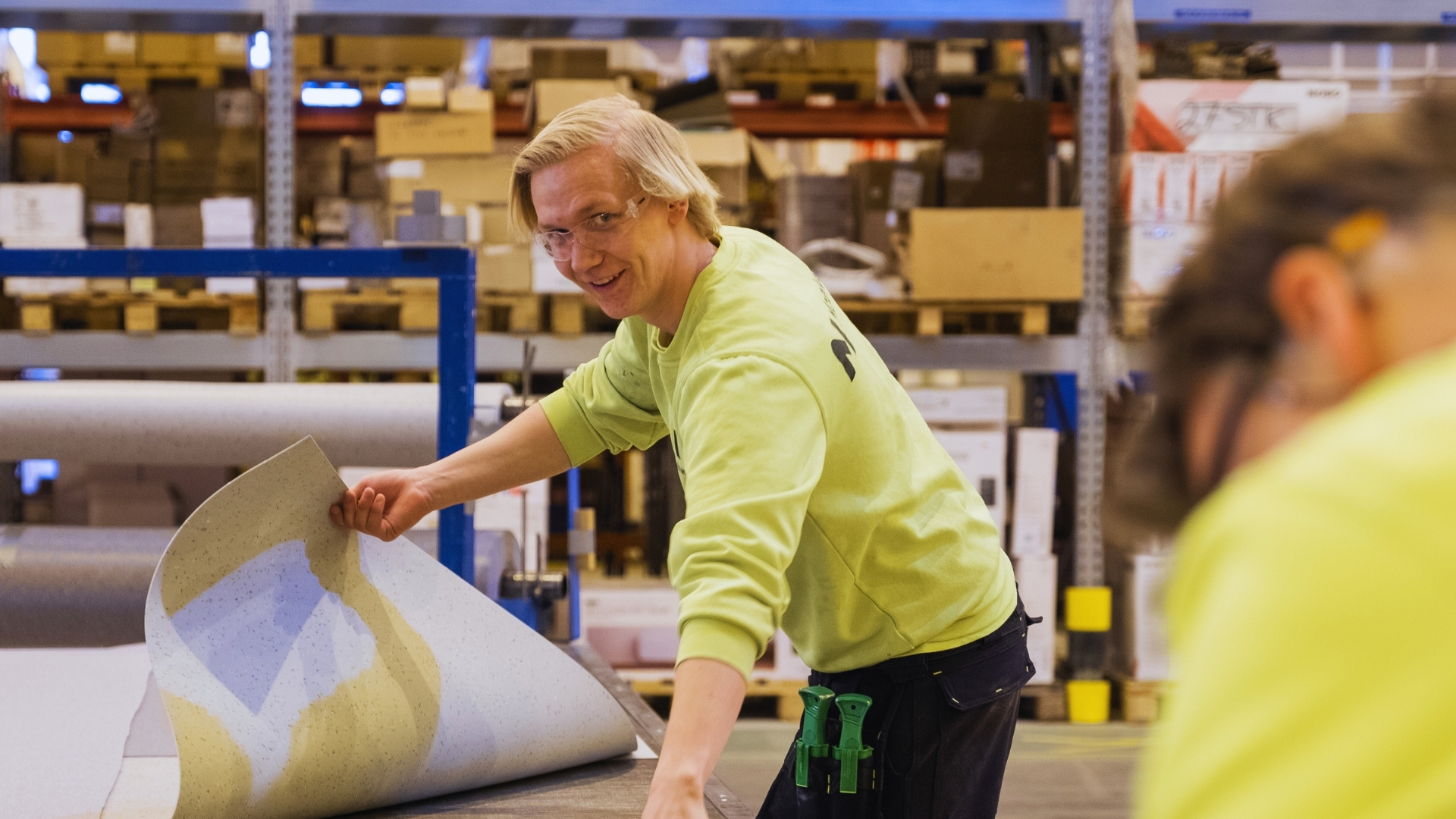 Робітник фабрики стоїть з підлоговим покриттям і усміхається в камеру.