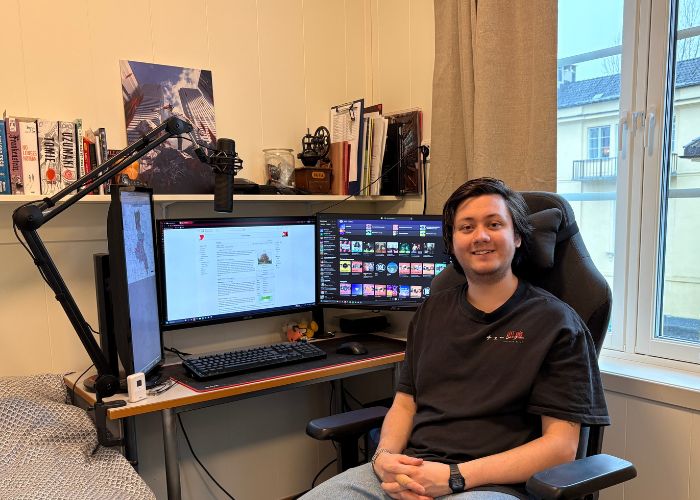 Person sitter ved en PC‑pult med to skjermer og mikrofon i et soverom.