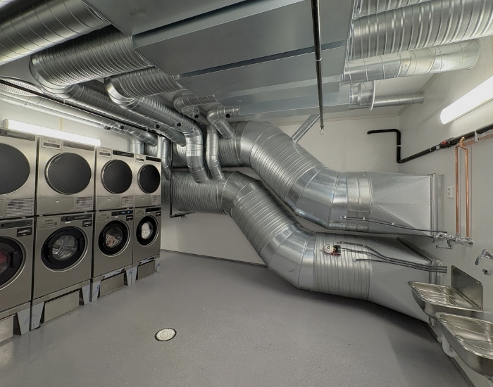 Moderne vaskerom med industrielle vaskemaskiner og tørketromler, synlige ventilasjonsrør og to utslagsvasker langs veggen.
