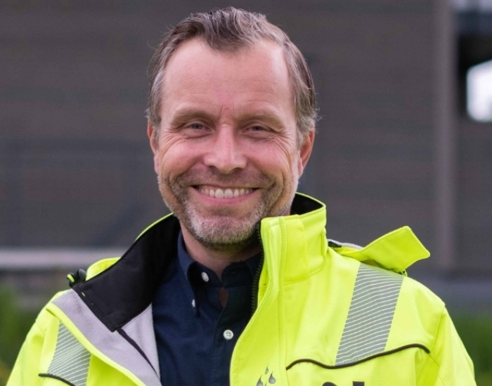 Smilende mann i synlighetsjakke utenfor en bygning.