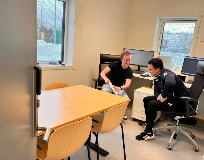 To smilende personer som sitter på sine kontorplasser lener seg mot hverandre.