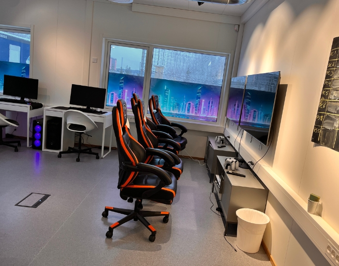 Rom med gamingstoler, skjermer og folierte vinduer med skyskrapermotiv.