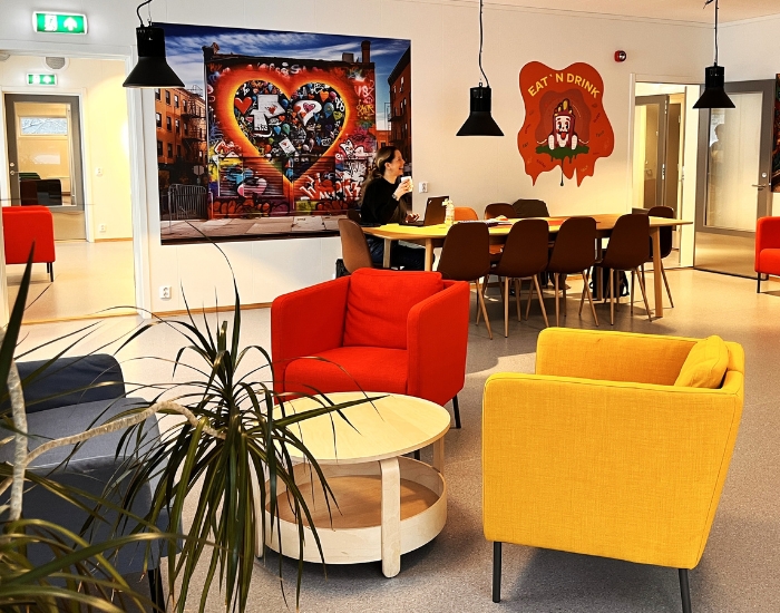 En rød og en gul stol rundt et hvitt bord. Smilende person som sitter på langbord bak.