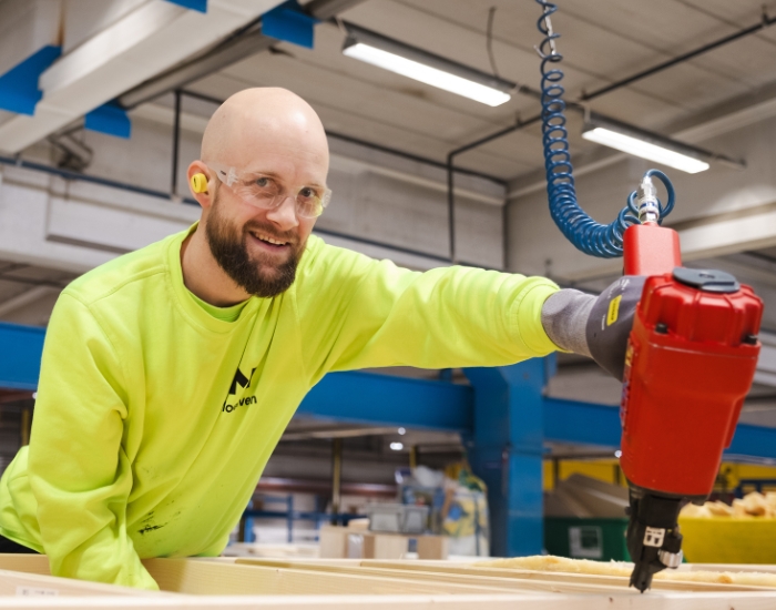 Smilende fabrikkarbeider holder en spikerpistol mot et byggelement.
