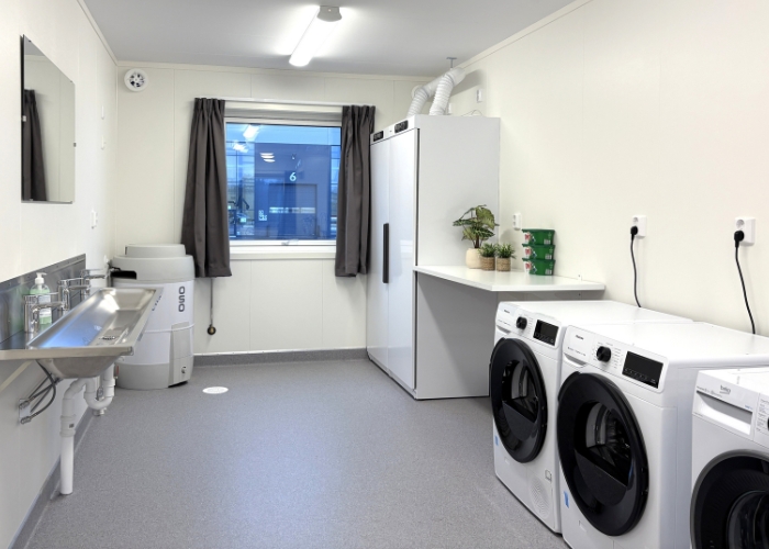 Felles vaskerom med stort vindu, vaskerenne, vaskemaskiner og tørkeskap i boligrigg.