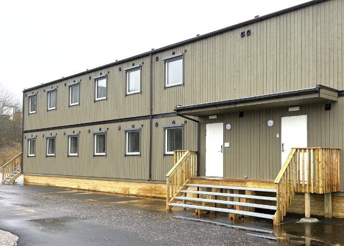 Inngangsparti til modulbygg med trapp og to inngangsdører. Brungrønt bygg med sorte eksteriørdetaljer.