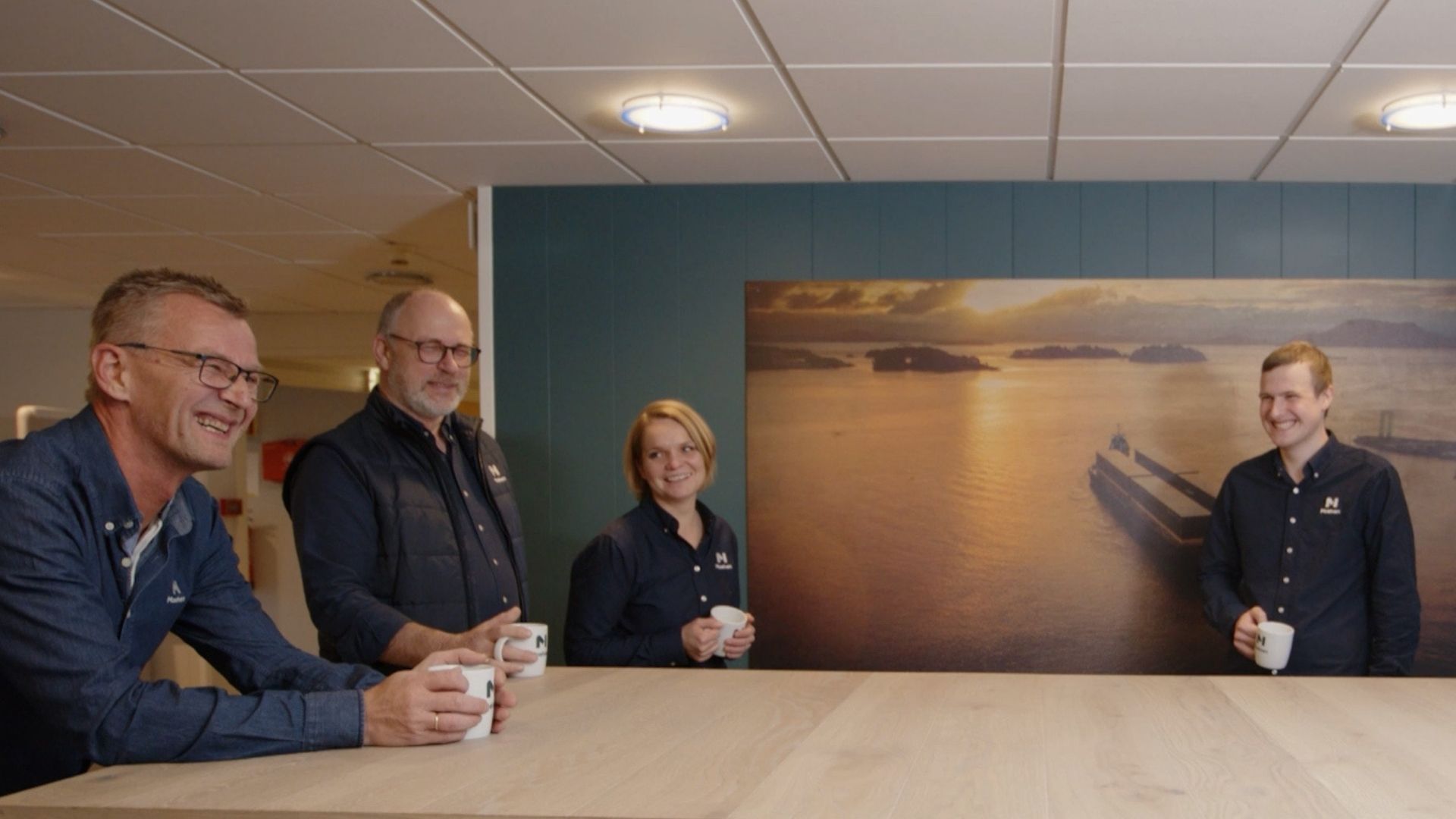Fire leende personer med kaffekopp står inntil et stort bord.