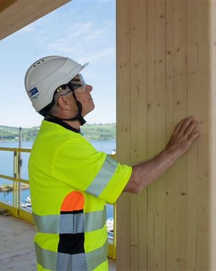 Person med hjälm och signalkläder som har handen på en limträbalk av ljust trä