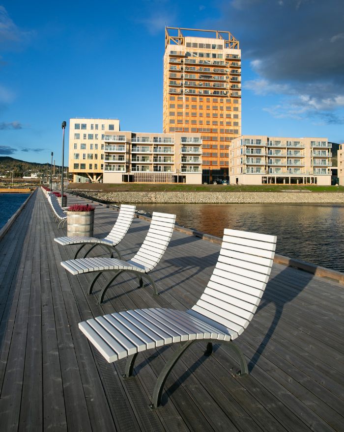Mjöstårnet sett från bryggan i solljus med vita solstolar framför.
