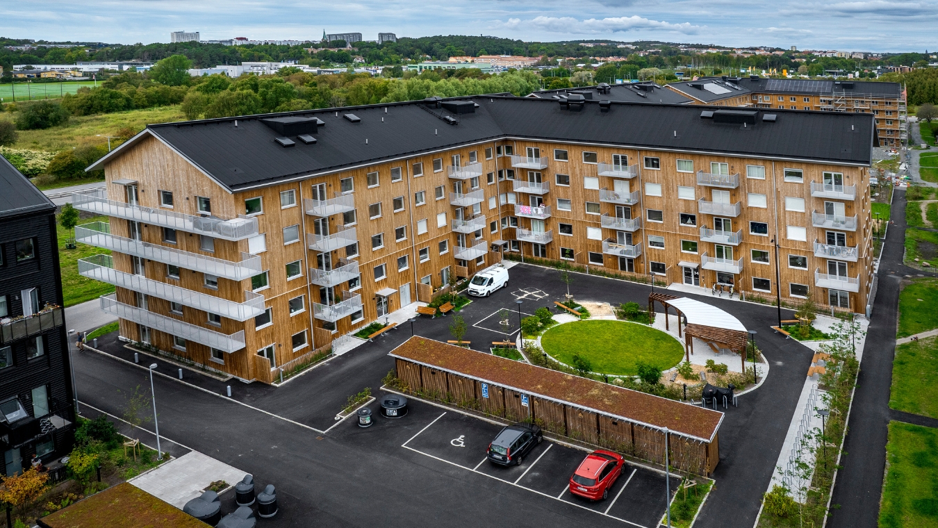 Översiktsbild över bostadshus sett utifrån med träfasad, parkering på innergård
