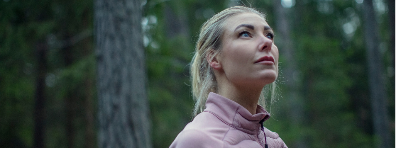 Kvinna i skogen som tittar upp mot himlen/topparna av träden