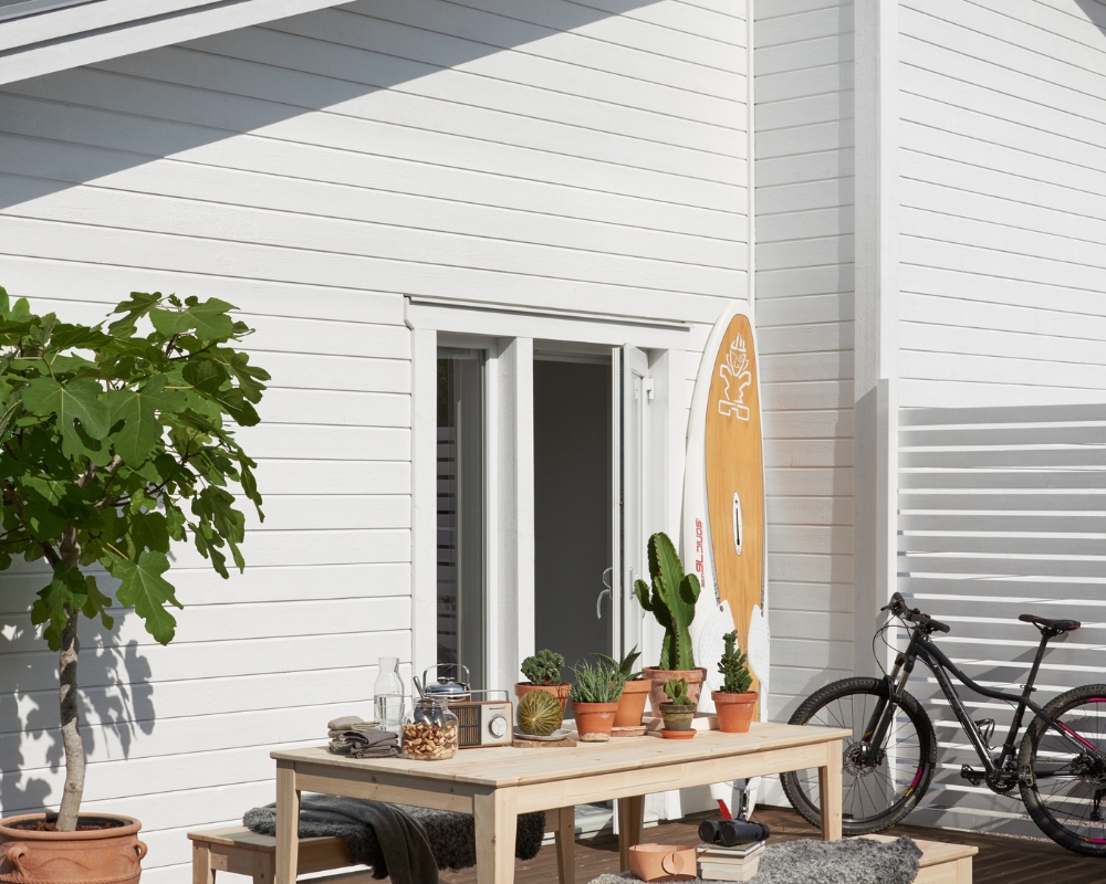 Vit ytterpanel och träbord med bänkar, växter och en cykel lutad mot ett staket