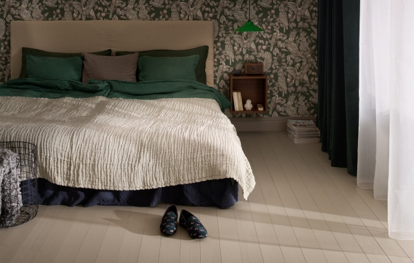 Ett sovrum med målat trägolv, en säng, gardiner, sängbord, en vägg med en mönstrad tapet och övriga inredningsdetaljer