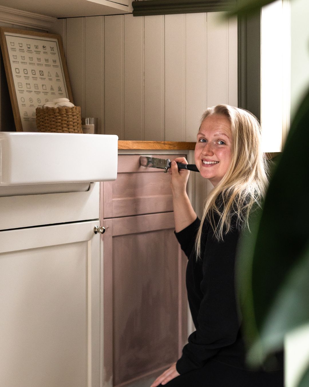 En ljushårig kvinna sitter på golvet i en tvättstuga, ler mot kameran, håller i en pensel och målar ett vitt skåp rosar