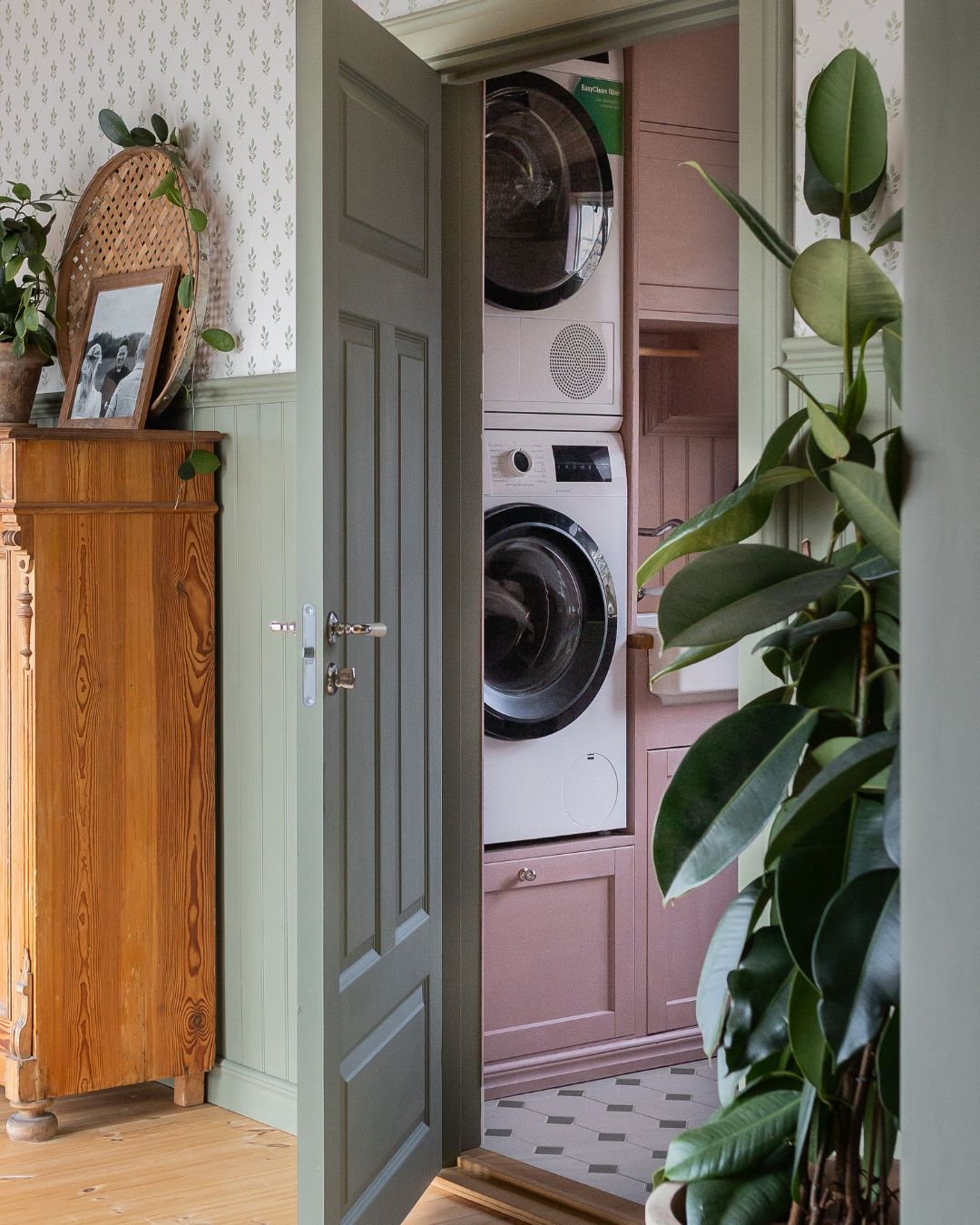 Hall med grönmålad pärlspont och öppen dörr till en tvättstuga. Innanför dörren syns staplad tvättmaskin och torktumlare, rosa skåp och ett tvättställ, medan en stor grön krukväxt står i förgrunden.