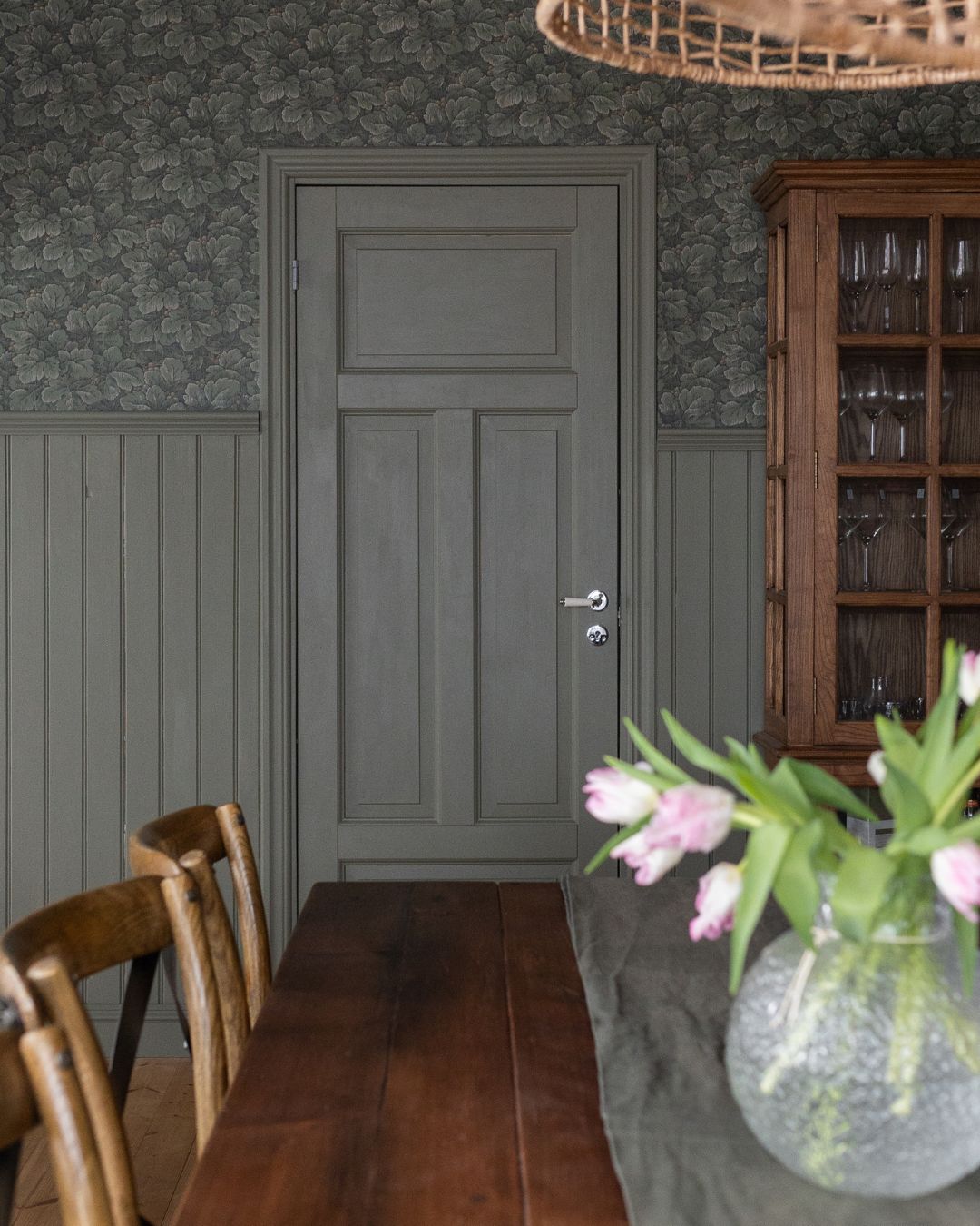 Matsal med grön bröstpanel av pärlspont och mörk blommönstrad tapet ovanför. Ett långt träbord med stolar står i förgrunden, och längs väggen syns en dörr  i samma kulör som pärlsponten samt ett vitrinskåp i trä med glasdörrar.