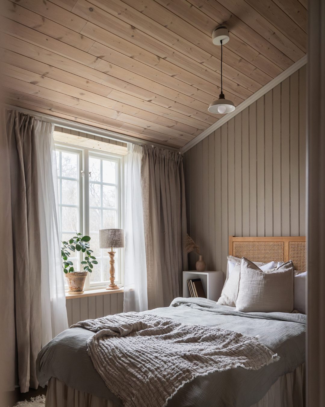 Sovrum i beige toner, sänggavel i rotting och stående pärlspont på väggarna och vitvaxat trätak.