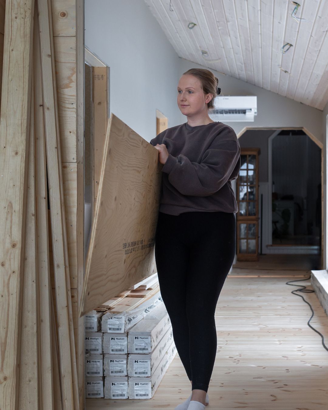 En kvinna bär en plywoodskiva i ett hus under renovering. Längs väggen står staplade paket med träpanel, och rummet har ljust trägolv, vitmålat trätak och väggar som delvis är under uppbyggnad.