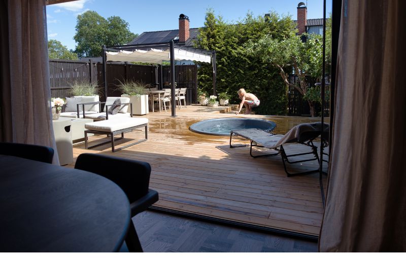 Utsikt över en altan med pergola och barn som hoppar i en liten pool sett inifrån huset