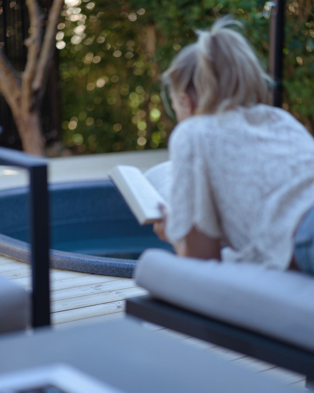Kvinna ligger och läser en bok utomhus och en liten pool syns längre bort.