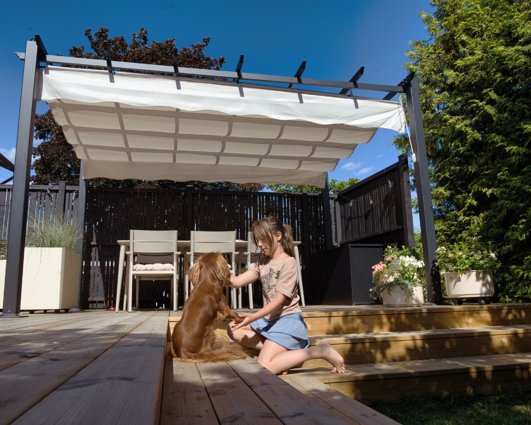 En flicka leker med en hund på en altan med trappor i bakgrunden en pergola med vitt tak