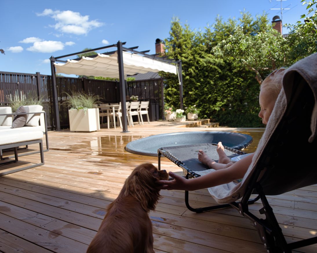 Barn i solsäng klappar en hund som sitter på en altan som är blöt vatten från en rund pool