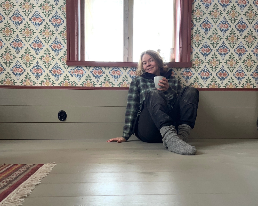 En kvinna sitter på ett gråmålat trägolv under ett fönster