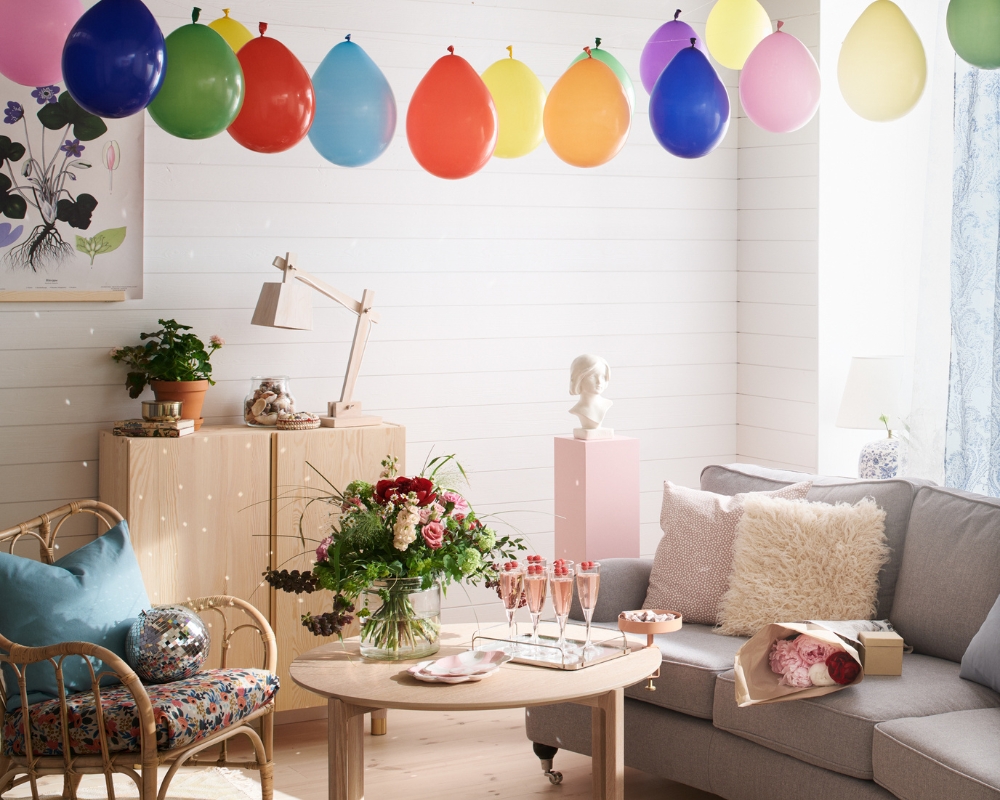 Ett vardagsrum med trägolv, vit träpanel på väggen och ett tak med panel och bjälkar, en grå soffa, fåtölj, ett ljust träbord och en byrå, samt  övriga inredningsdetaljer som tex ballonger