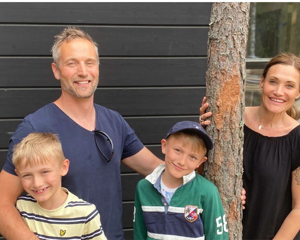 En familj med en man, en kvinna och två pojkar, framför ett hus med svart ytterpanel och ett träd