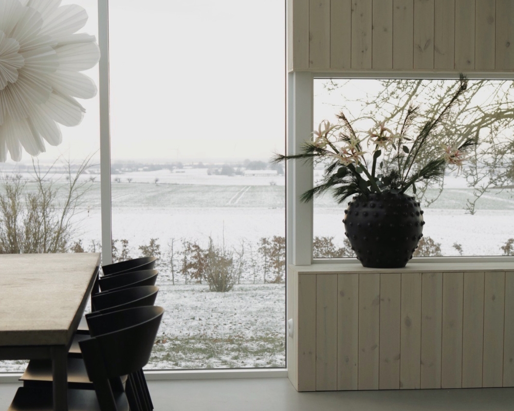Bord och stolar, inglasad dörr med utsikt mot en åker med snö, samt en stor svart vas med blommor, och en hängande stjärna vid inglasade dörren