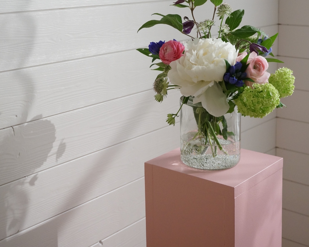 En rosa piedestal och en vas med blommor, i bakgrunden en vit träpanel