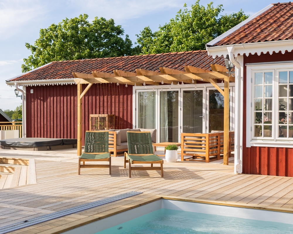 Ett rött hus med en altan av trä, en pool, jacuzzi och utemöbler