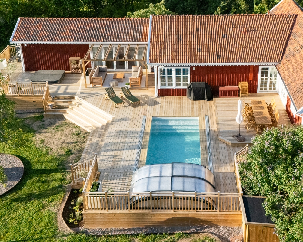 Ett rött hus och en stor altan i trä, pool, jacuzzi och utomhusmöbler, samt en tomt med gräs och träd