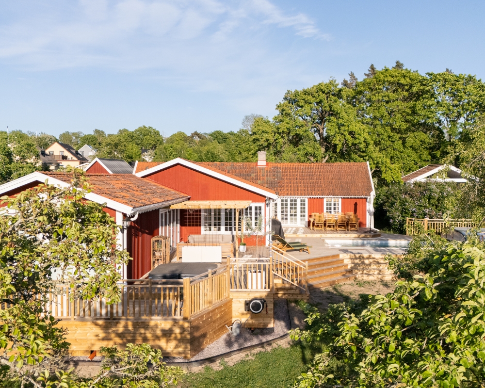 Ett rött hus, altan i trä, pool. utemöbler, tomt med träd