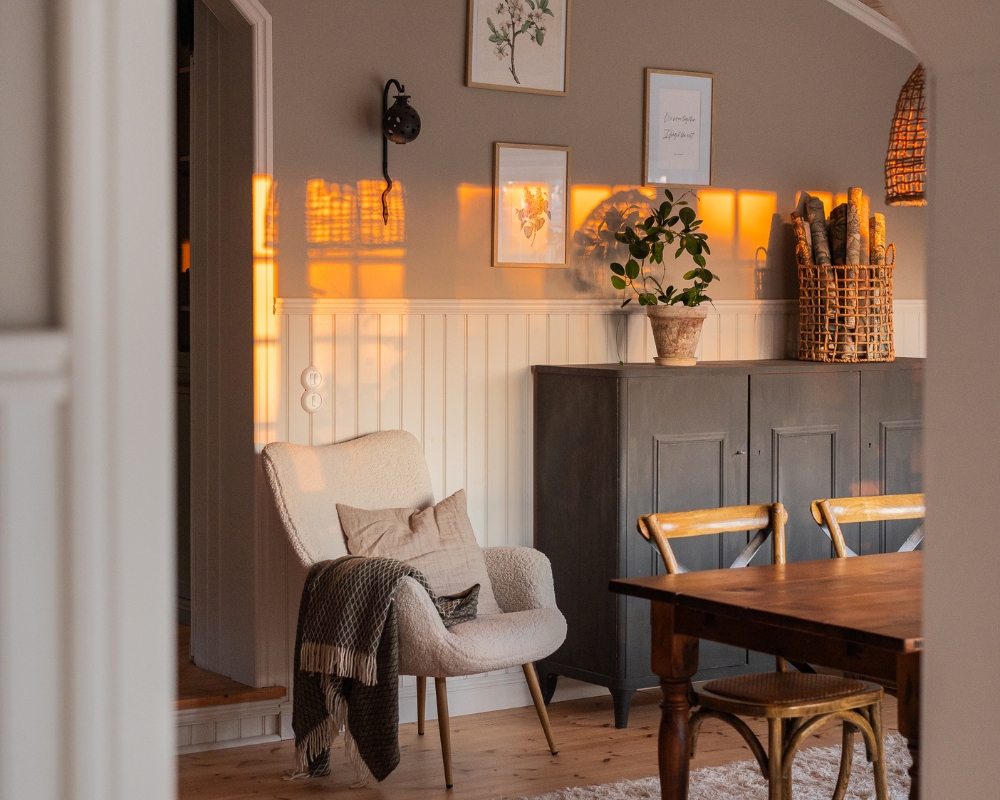 Matsal med vit bröstpanel i pärlspont. Ett brunt matbord och stolar i trä. En fåtölj teddypläd  och ett grått skåp, samt dekorationer