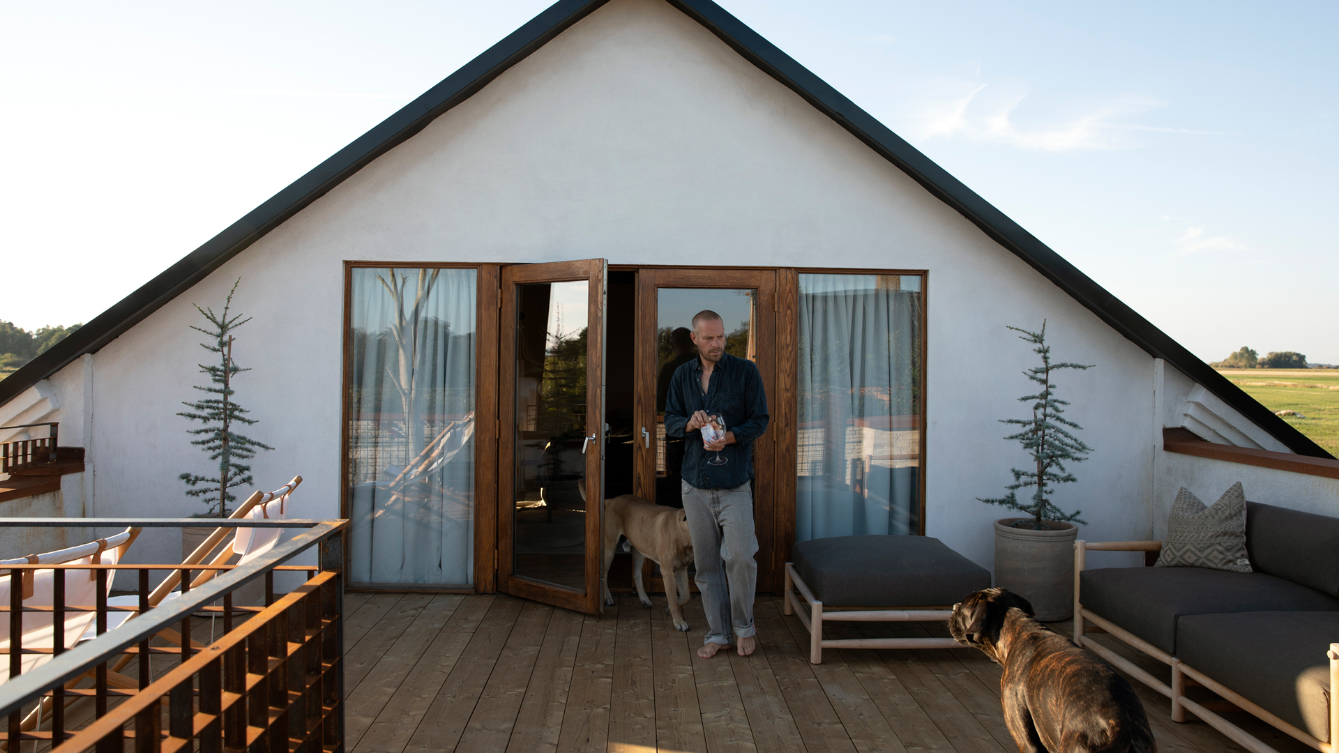 Gustav Ovland står barfota på takterrassen i TermoGran tillsammans med två hundar, framför dubbelglasdörrarna till ovanvåningen. På sidorna syns loungemöbler och två planterade små barrträd.