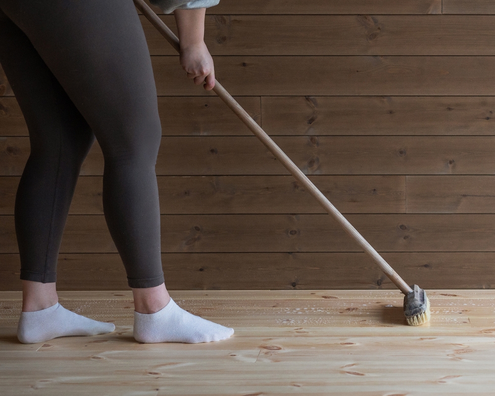 Benen av en kvinna syns medans hon rengör ett trägolv med en borste