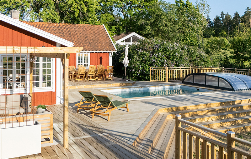 Ett trött hus med en stor altan i trä, och en pool, samt utemöbler och skog